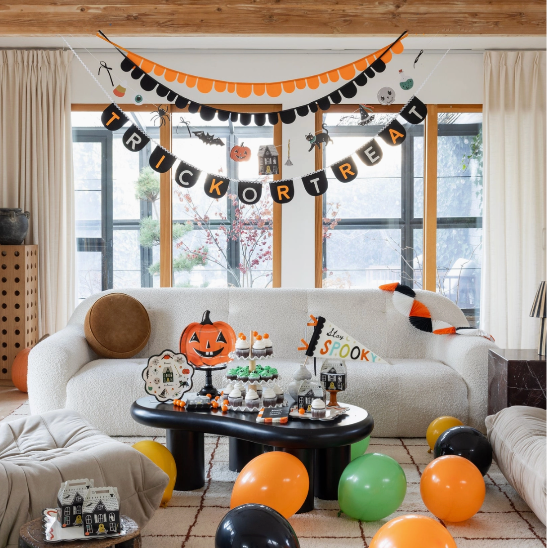 A styled living room decorated for Halloween, featuring a black and orange 'Trick or Treat' banner, a 'Happy St. Patrick's Day' banner, pumpkins, balloons, and a decorated white sofa.