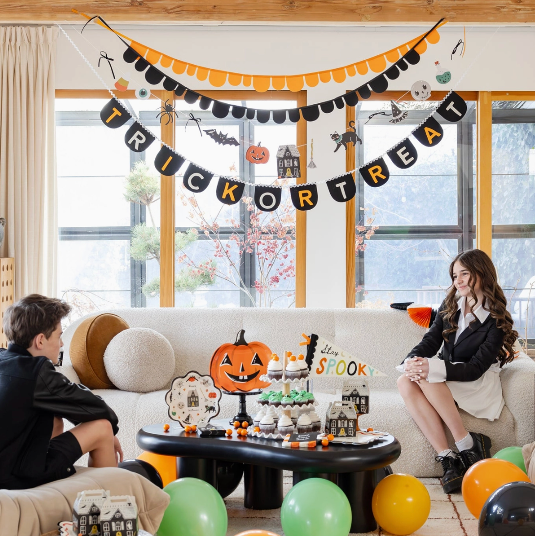 A Halloween-themed party setup in a bright living room with two children seated on a white sofa. The room is decorated with a black and orange 'Trick or Treat' banner, a pumpkin garland, spiderwebs, and a coffee table covered in fall decorations, including a jack-o'-lantern, cupcakes, and a 'The Best of Halloween' sign.
